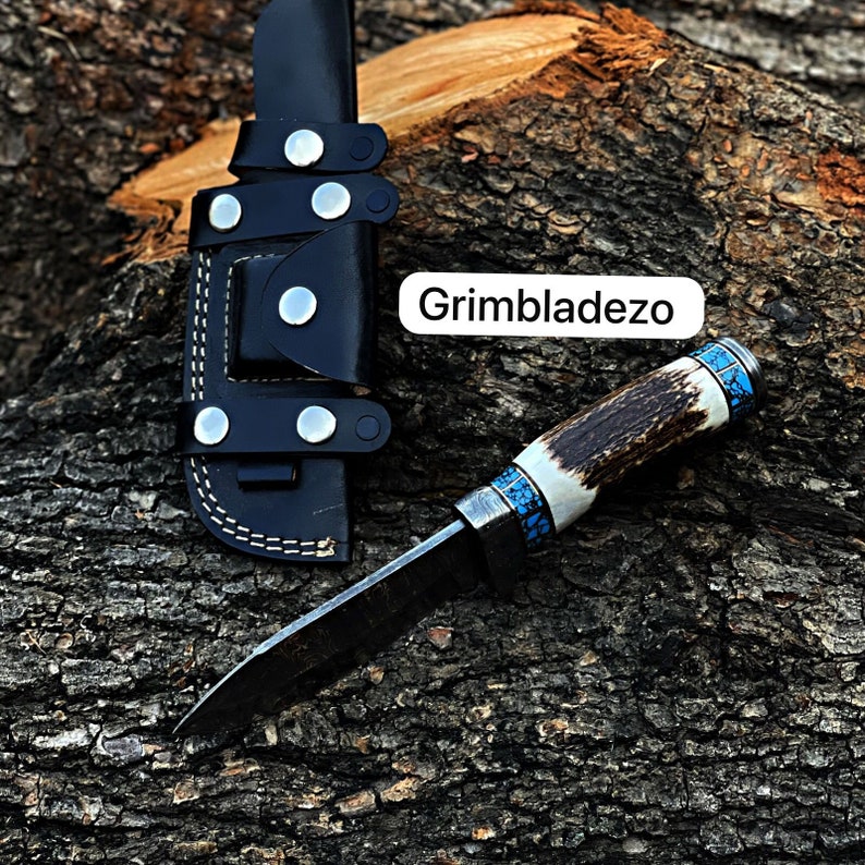 Może przedstawiać: Czarna sk&oacute;rzana pochwa z nożem o rękojeści z poroża jelenia i niebiesko-turkusową inkrustacją. N&oacute;ż leży na drewnianej powierzchni. W obrazie widoczny jest tekst "Grimbladezo".