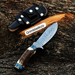 Może przedstawiać: N&oacute;ż myśliwski z ostrzem ze stali damasceńskiej i rękojeścią z poroża jelenia. Rękojeść zdobiona jest niebieskimi turkusowymi intarsjami. N&oacute;ż znajduje się w czarnej sk&oacute;rzanej pochwie.