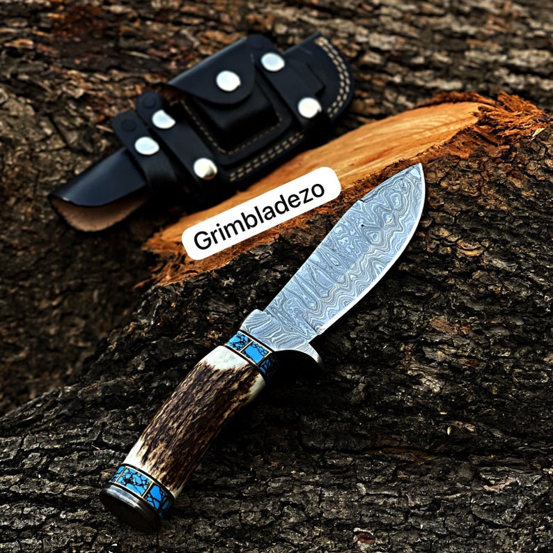 Może przedstawiać: N&oacute;ż myśliwski z ostrzem ze stali damasceńskiej i rękojeścią z poroża jelenia. Rękojeść zdobiona jest niebieskimi turkusowymi intarsjami. N&oacute;ż znajduje się w czarnej sk&oacute;rzanej pochwie.