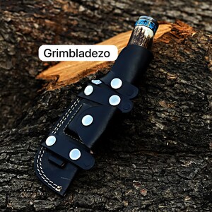 Może przedstawiać: Czarna sk&oacute;rzana pochwa na n&oacute;ż z brązową i niebieską rękojeścią. Pochwa ma białe szwy i srebrne zatrzaski. W obrazie widoczny jest tekst "Grimbladezo".