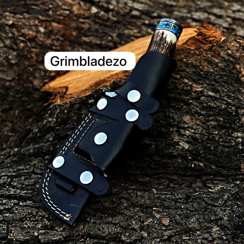 Może przedstawiać: Czarna sk&oacute;rzana pochwa na n&oacute;ż z brązową i niebieską rękojeścią. Pochwa ma białe szwy i srebrne zatrzaski. W obrazie widoczny jest tekst "Grimbladezo".