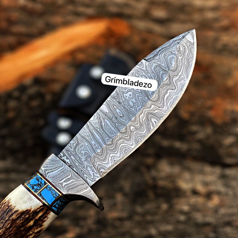 Może przedstawiać: Zbliżenie na n&oacute;ż myśliwski z ostrzem ze stali damasceńskiej i rękojeścią z poroża jelenia. Ostrze ma niebieską i turkusową inkrustację w pobliżu rękojeści. Na ostrzu widoczny jest napis "Grimbladezo".