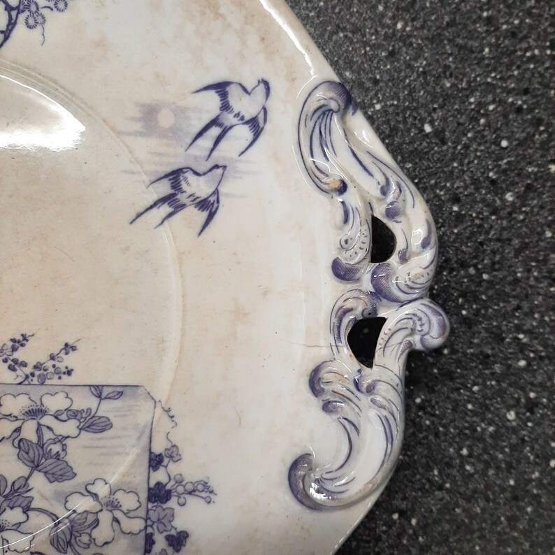 Puede incluir: Un plato de servir de cer&aacute;mica blanca con dise&ntilde;os florales y de p&aacute;jaros azules. El plato tiene dos asas ornamentadas a los lados.