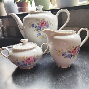 Puede incluir: Un juego de tres tazas de té de porcelana blanca con diseños florales. La tetera tiene un asa y un pico dorados, mientras que la jarra de crema y el azucarero tienen bordes dorados. Los diseños florales presentan flores rosas, azules y amarillas.