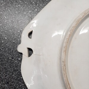 Puede incluir: Plato de cer&aacute;mica blanco con borde festoneado y dos asas recortadas. El plato tiene una peque&ntilde;a grieta en el borde.