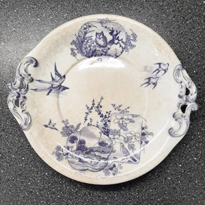Puede incluir: Un plato de cer&aacute;mica blanco con un dise&ntilde;o floral azul y blanco. El plato presenta un dise&ntilde;o central de p&aacute;jaros y flores, con un borde de p&aacute;jaros y hojas. El plato tiene un borde festoneado y dos asas.