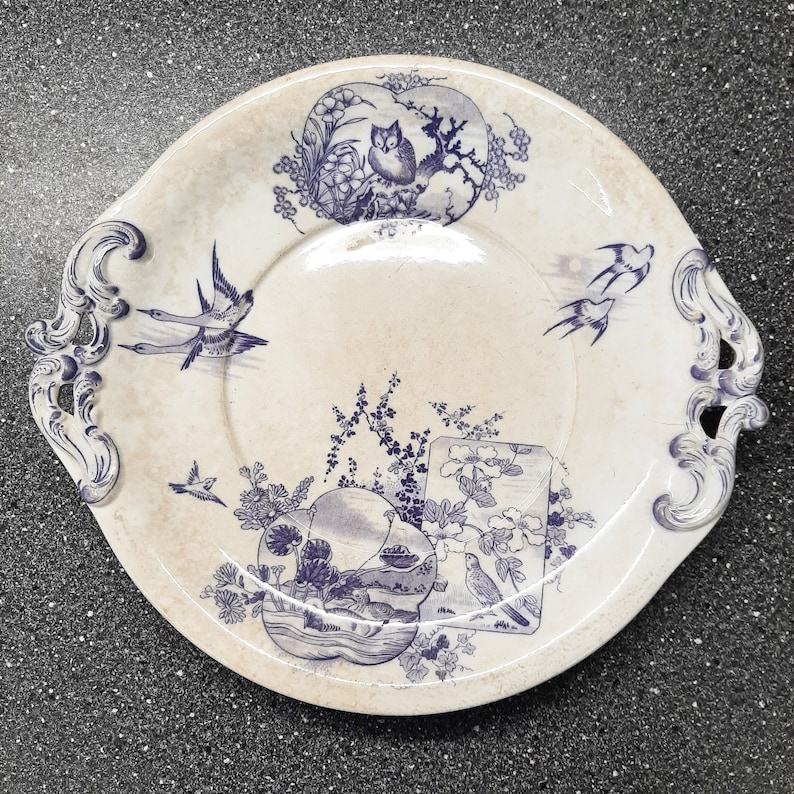 Puede incluir: Un plato de cer&aacute;mica blanco con un dise&ntilde;o floral azul y blanco. El plato presenta un dise&ntilde;o central de p&aacute;jaros y flores, con un borde de p&aacute;jaros y hojas. El plato tiene un borde festoneado y dos asas.