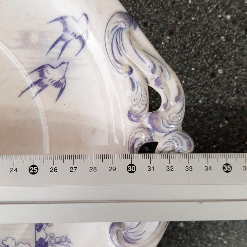 Puede incluir: Un plato de servir de cer&aacute;mica blanca con un dise&ntilde;o floral azul y morado y dos p&aacute;jaros azules. El plato tiene un asa en el lateral y mide aproximadamente 76 cm de largo.