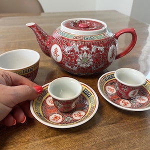 Könnte beinhalten: Ein rotes und weißes Keramik-Teeservice mit einer Teekanne, zwei kleinen Tassen und zwei Untertassen. Die Teekanne und die Tassen weisen ein Blumen- und geometrisches Muster mit chinesischen Schriftzeichen auf. Das Set steht auf einer Holzoberfläche.