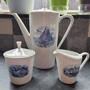 Op de afbeelding: Een witte porseleinen koffiekan, roomkan en suikerpot met blauwe en witte nautische ontwerpen. De koffiekan heeft een hoog, cilindrisch lichaam met een tuit en een handvat. De roomkan en suikerpot zijn kleiner en hebben ronde lichamen met deksels. Alle drie de stukken hebben een blauw en wit zeilbootontwerp met een blauw en wit huisontwerp op de suikerpot.