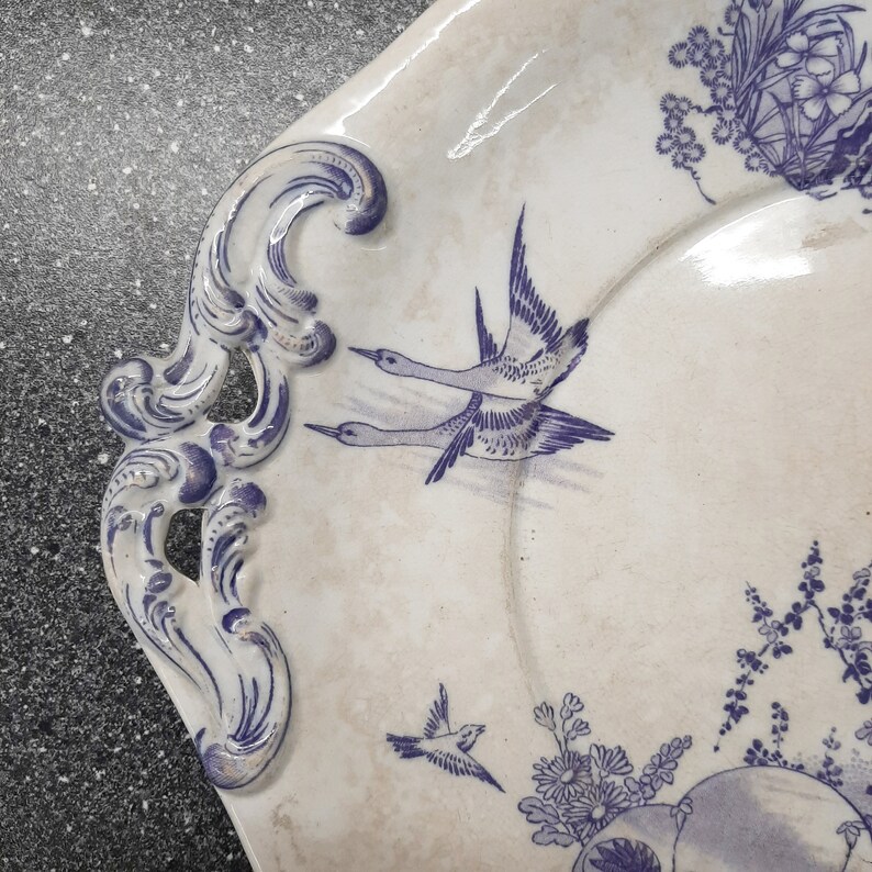 Puede incluir: Un plato de servir de cer&aacute;mica blanca con un dise&ntilde;o floral azul y blanco. El plato presenta dos p&aacute;jaros en vuelo y un patr&oacute;n floral alrededor del borde.
