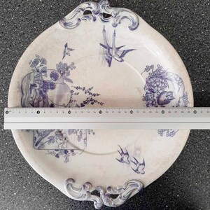 Puede incluir: Un plato de servir de cer&aacute;mica blanca con un dise&ntilde;o floral azul y blanco. El plato tiene dos asas y un borde festoneado. El dise&ntilde;o presenta p&aacute;jaros, flores y hojas.