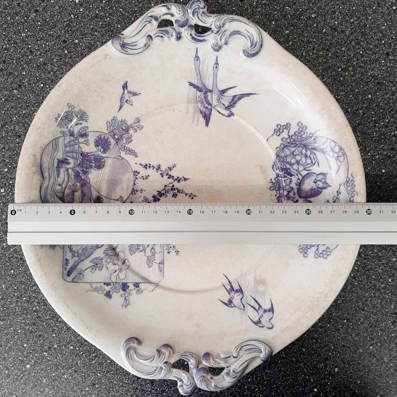 Puede incluir: Un plato de servir de cer&aacute;mica blanca con un dise&ntilde;o floral azul y blanco. El plato tiene dos asas y un borde festoneado. El dise&ntilde;o presenta p&aacute;jaros, flores y hojas.