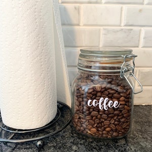 May include: A glass jar filled with roasted coffee beans, labeled with the word "coffee" in white script. A roll of white paper towels sits to the left. The jar has a metal clasp and a glass lid.