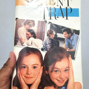 Puede incluir: Una cinta de VHS de la película de Disney "Juego de Gemelas". La portada presenta imágenes de los actores de la película, incluidas dos niñas y otros actores. El título está en letras grandes y en negrita con una franja azul.