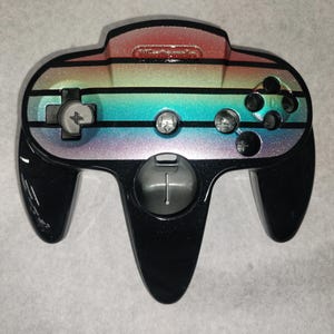 May include: A black Nintendo 64 controller with a rainbow striped design across the top. The controller features a gray directional pad, gray buttons, and a gray analog stick. The Nintendo logo is visible at the top.