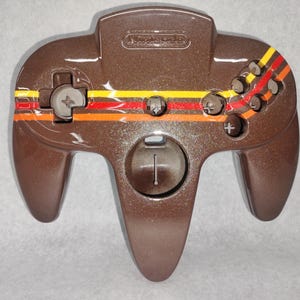 May include: A brown Nintendo 64 controller with a retro stripe design in orange, yellow, and red. The controller features a directional pad, analog stick, and multiple buttons, with the Nintendo logo at the top.