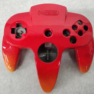 May include: A red and orange gradient Nintendo 64 controller. The controller has a directional pad, analog stick, and buttons. The word "Nintendo" is embossed on the top.