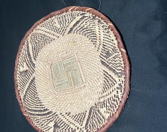 May include: A round, hand-woven basket tray featuring a central square pattern of light tan reeds surrounded by concentric circles of alternating dark brown and light tan geometric designs, finished with a thick, braided dark brown rim.