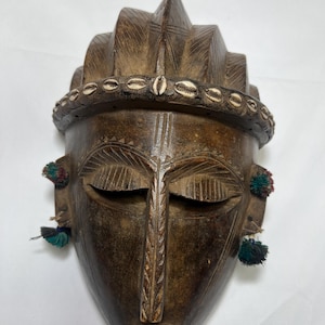 Puede incluir: Una máscara de madera marrón oscuro con una cara tallada y una corona de conchas. La máscara tiene los ojos cerrados y una pequeña boca. Hay borlas verdes y rojas que cuelgan de las orejas.