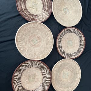 Puede incluir: Colección de seis cestas redondas tejidas. Las cestas presentan tonos beige y marrón naturales, algunas con intrincados patrones geométricos en los bordes. Las cestas varían en tamaño y diseño, mostrando una estética rústica y artesanal.
