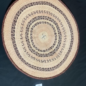 Puede incluir: Una cesta redonda y plana tejida con círculos concéntricos de fibras naturales en color beige y marrón oscuro. El diseño incluye bandas alternas de beige sólido y patrones geométricos complejos en marrón oscuro. Se ve una pequeña etiqueta blanca con el número #1.