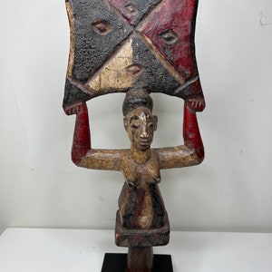 May include: A wooden sculpture of a woman holding a large, painted, geometric shape above her head. The sculpture is painted in shades of red, black, and white.