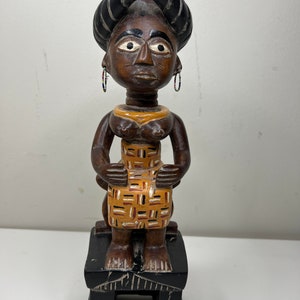 以下が含まれることがあります： 黒いターバンをかぶった女性の、黄色とオレンジの模様のドレスを着た木製の置物。置物は黒い木製の台座の上に立っている。