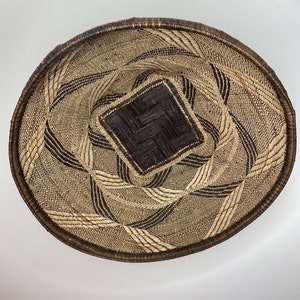 Puede incluir: Una cesta redonda tejida con un diseño cuadrado central. La cesta está hecha de fibras naturales marrones y beige, con detalles en marrón oscuro. El borde es de un marrón más oscuro y el tejido forma un patrón geométrico.