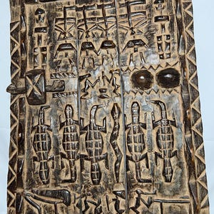 Pode incluir: Painel de madeira entalhada com intrincados desenhos em relevo. O painel apresenta padrões geométricos, figuras estilizadas e um mecanismo central de travamento. A madeira tem uma aparência escura e envelhecida, com grão e textura visíveis.