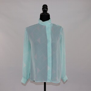 May include: A light blue blouse with a high collar and long sleeves. The sheer fabric features a subtle geometric pattern. The blouse is displayed on a black mannequin.