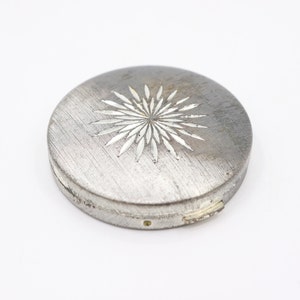 May include: A silver compact with a starburst design. The compact has a brushed metal finish and a hinged lid.