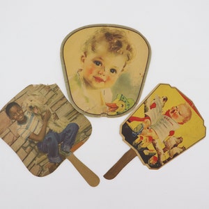 May include: Three vintage paper hand fans with colourful illustrations. One fan features a young boy with a dog, another features a baby with a duck, and the third features a young girl with a teddy bear.