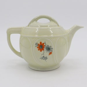 Peut inclure: Une théière en céramique de couleur crème avec un motif floral. La théière a un corps rond avec un motif cannelé et un couvercle avec une poignée. Le motif floral présente des fleurs orange et grises.