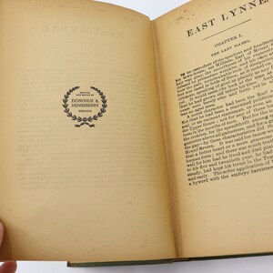 May include: An open book with a cream-colored cover and yellowed pages. The book is titled "East Lynne" and the text on the page describes the Earl of Mount Severn. The book has a black and white publisher's logo on the left page.