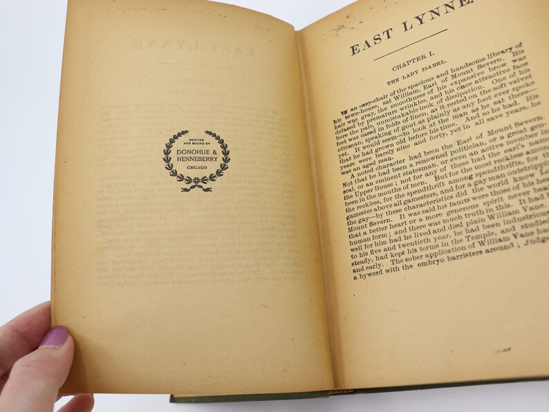May include: An open book with a cream-colored cover and yellowed pages. The book is titled "East Lynne" and the text on the page describes the Earl of Mount Severn. The book has a black and white publisher's logo on the left page.