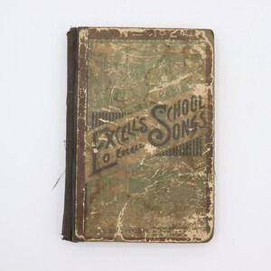 May include: A vintage book titled "Excel's School Songs" with a worn, green cover and brown spine. The book is slightly damaged and has a frayed edge.
