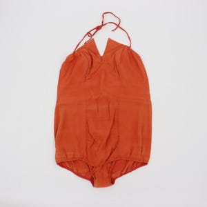 May include: A burnt orange one-piece swimsuit with a halter neck and a low back. The swimsuit has a V-neckline and a red trim around the leg openings. The fabric appears to be a lightweight material.