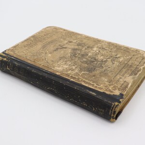 May include: A vintage, black-bound book with a worn, tan cover. The cover features a faded illustration and text that reads "The Land and the Book".