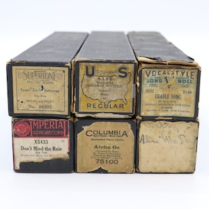 May include: Six vintage music player boxes with labels. The boxes are black with white and gold lettering. The labels include the titles "Sweet Little Buttercup", "Wailana Drowsy Waters", "Cradle Song", "Don't Mind the Rain", "Aloha Oe", and "Aline the".