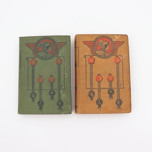 May include: Two vintage hardcover books with illustrated covers. The covers feature a woman in a crown with wings and a banner with stars. One book has a green cover and the other has a brown cover.