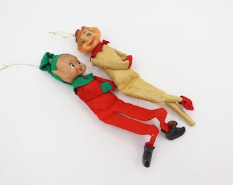 May include: Two vintage Christmas elf ornaments. One elf is dressed in red with a green hat and collar, the other in gold. Both have painted faces, black shoes, and red accents.