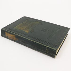 May include: A vintage, dark green book with gold lettering and a decorative gold emblem on the cover. The book's spine shows signs of wear, suggesting its age. The book is set against a plain white background.