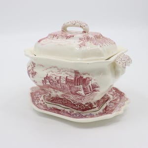 May include: A white ceramic soup tureen with a pink and white floral pattern. The tureen has a lid with a handle and a base with a matching plate. The tureen features a scene of a castle in the countryside.
