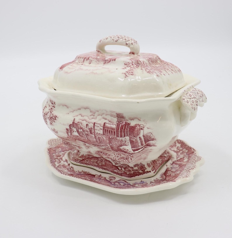 May include: A white ceramic soup tureen with a pink and white floral pattern. The tureen has a lid with a handle and a base with a matching plate. The tureen features a scene of a castle in the countryside.