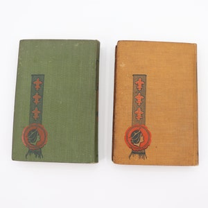 May include: Two vintage hardcover books with green and brown covers. Both books feature a decorative design with a woman's profile and a fleur-de-lis motif.