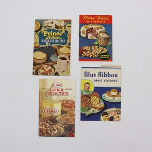 May include: Four vintage recipe booklets are displayed. The booklets feature titles such as "Prince Golden Macaroni Recipes," "Menu Magic in a Nutshell," "Crisco Cookie Collection," and "Blue Ribbon Malt Extract." The covers showcase illustrations of food.