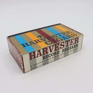 May include: A box of Harvester Record Breaker cigars. The box is brown with a multi-colored stripe design. The text on the box reads "Harvester Record Breaker A Quality Cigar Named After A Champion."