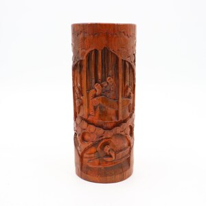 May include: A cylindrical wooden vase with intricate carvings depicting a scene of people and trees. The vase is made of dark brown wood and has a smooth, polished finish.