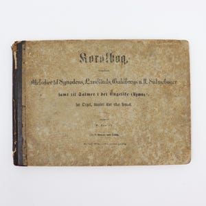 May include: A vintage book titled "Koralbog" with the text "Melodier til Synodens, Landstads, Guldbergs o.fl. Salmebøger samt til Salmer i det Engelife (Hymns), for Orgel, Mandt eller bas" printed on the cover.